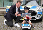 Staatssekretär Sandro Kirchner neben Kind mit Polizeikappe auf Polizeibobbycar vor Polizei-MINI