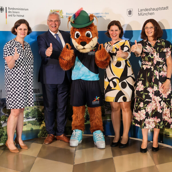 Gruppenbild mit Daumen hoch: Staatssekretärin des Bundesinnenministeriums Juliane Seifert, Bayerischer Innenminister Joachim Herrmann, Maskottchen „Gfreidi“, 3. Bürgermeisterin der Landeshauptstadt München Verena Dietl und Geschäftsführerin Olympiapark München Marion Schöne