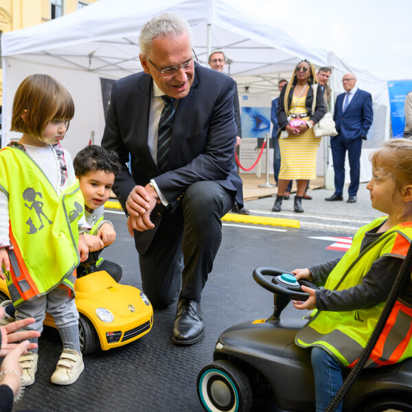 Innenminister Joachim Herrmann kniet zu Kindern in Verkehrswarnfesten auf Bobycars