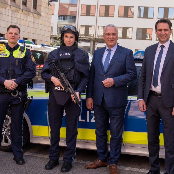 Innenminister Joachim Herrmann und Justizminister Georg Eisenreich neben Polizist und Polizistin in Schutzkleidung vor Polizeiauto
