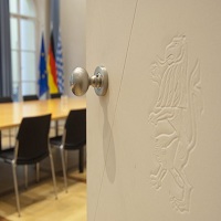 Das Foto zeigt einen Ausschnitt eines Sitzungssraums, im Hintergrund sind die Fahnen Bayerns, Deutschlands und die Europaflagge ersichtlich. 